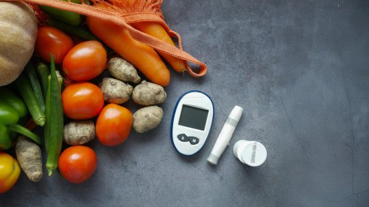 Blood sugar monitor and diabetic testing supplies next to fresh fruits, vegetables, and whole grains, symbolizing holistic blood sugar management.