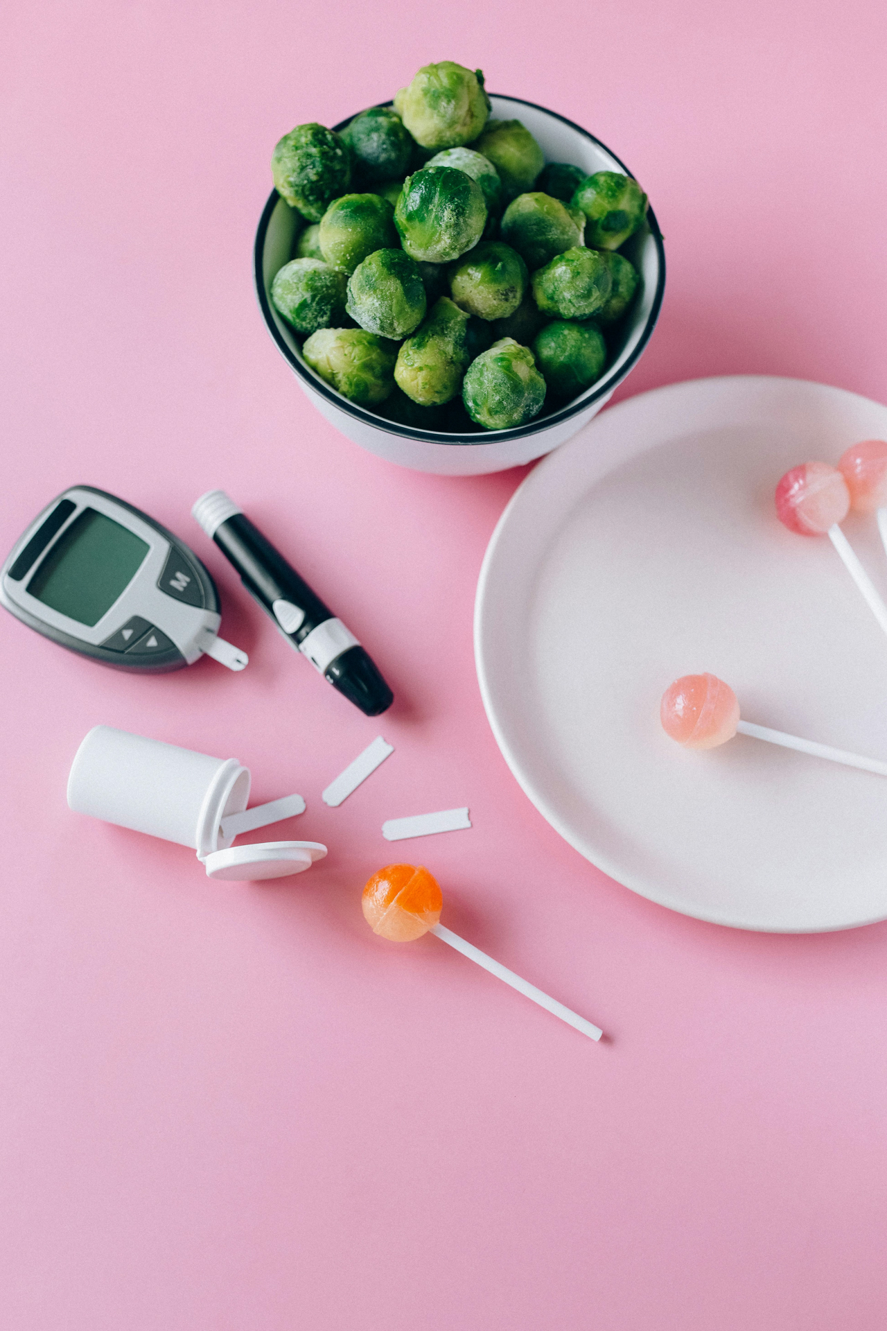 Best foods for managing blood sugar: bowl of fresh Brussels sprouts beside a glucose meter, test strips, lancet device, and contrasting sugar lollipops on a pink background.