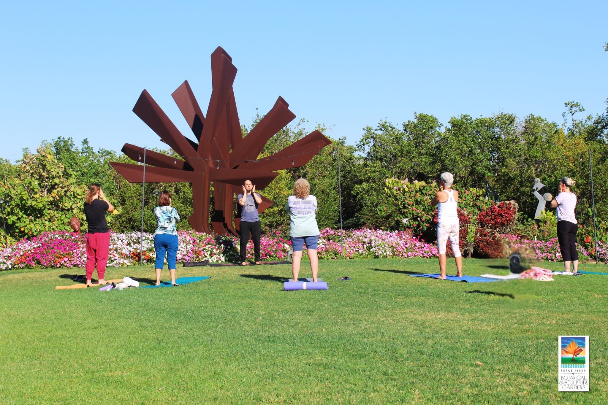 Group practicing Morning Activation at the Gardens near a large outdoor sculpture and vibrant flower beds at Peace River Botanical & Sculpture Gardens.